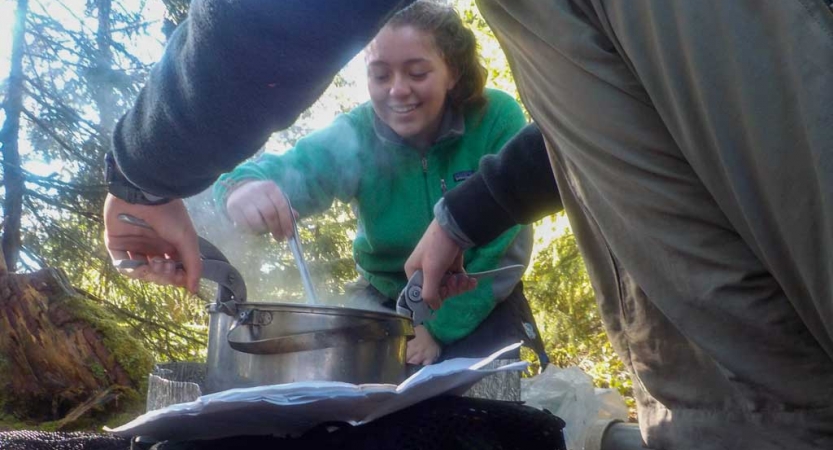 Students use kitchen supplies to cook in a wooded area. 